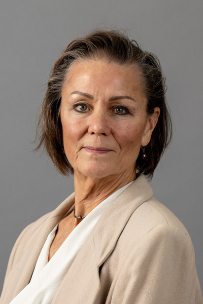 Portrait of a woman with short, dark brown hair, wearing a beige blazer, white blouse, and a simple necklace.